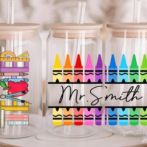 May include: Three clear glass tumblers with wooden lids and straws. The tumblers have colorful crayon designs and the text "Mr. Smith" on the middle tumbler.