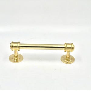 Puede incluir: Un tirador de armario dorado con una barra cilíndrica y placas de montaje circulares. El herraje tiene un acabado pulido y está diseñado para muebles. La longitud total es de aproximadamente 15 cm.