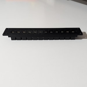 Peut inclure: Un composant électronique noir avec de multiples ouvertures et ports rectangulaires. Le composant est rectangulaire avec une série de petites fentes espacées uniformément le long du bord supérieur. Il comporte une série de ports rectangulaires le long du bord inférieur.