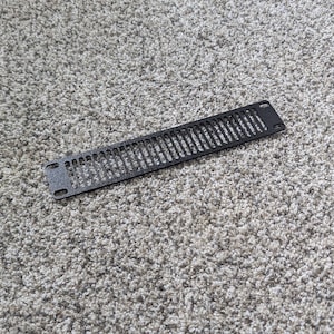 May include: A black, rectangular vent cover with a series of vertical slots. The vent cover is made of metal and has mounting holes on each end. It is lying on a gray, textured carpet.