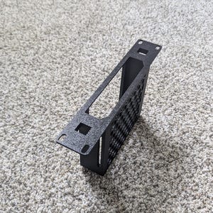 May include: A black metal bracket with a rectangular frame and a perforated side panel. The bracket has mounting holes and a textured finish. It is designed to hold electronic components.