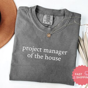 May include: A gray Comfort Colors t-shirt with the text "project manager of the house" in white. A gold necklace with a pendant rests on the shirt. A brown hat is visible in the upper left corner.
