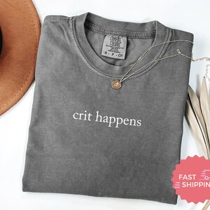 以下が含まれることがあります： 前面に白い文字で「crit happens」とプリントされたグレーのTシャツ。