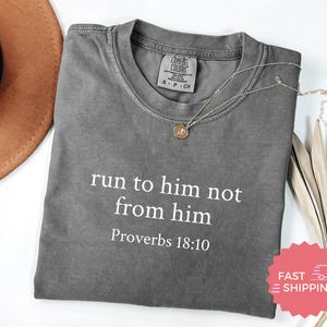 Puede incluir: Una camiseta gris con el texto "run to him not from him" y "Proverbios 18:10" impreso en blanco. Un collar dorado con un colgante está sobre la camiseta. Un sombrero marrón está en el fondo.