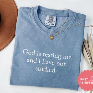 Könnte beinhalten: Ein hellblaues T-Shirt mit dem Aufdruck "God is testing me and I have not studied" in weiß.