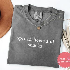 May include: A gray Comfort Colors t-shirt with the words "spreadsheets and snacks" in white. A gold necklace with a pendant rests on the shirt. A brown hat and dried plant are in the background.