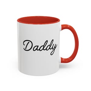 Puede incluir: Taza de cerámica blanca con borde y asa rojos. La taza tiene la palabra "Daddy" escrita en cursiva negra en la parte delantera.