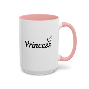 May include: White ceramic mug with a pink rim and handle. The mug has a black inscription that reads "Princess" with a heart and crown above the word.
