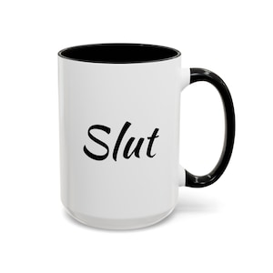 Könnte beinhalten: Weiße Keramiktasse mit schwarzem Rand und Henkel. Die Tasse hat das Wort "Slut" in schwarzer Kursivschrift auf der Vorderseite.