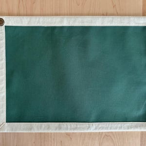 May include: A rectangular, dark green canvas with a cream-colored border. The canvas is framed with a cream-colored fabric and has brass grommets in each corner. The canvas is displayed against a light wood background.