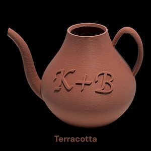Pode incluir: Um regador de cor terracota com superfície texturizada, apresentando um bico curvo e uma alça. As letras K + B estão em relevo na parte frontal do corpo arredondado. A palavra Terracotta está impressa em tom castanho claro abaixo.