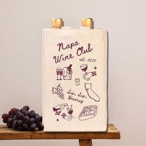 May include: A white canvas wine tote bag with the text "Napa Wine Club est. 2024" and illustrations of wine bottles, grapes, glasses, and a map of California. The bag has two pockets on the sides for wine bottles.