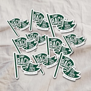 Puede incluir: Un conjunto de pegatinas verdes y blancas con forma de bandera. Cada pegatina presenta el texto "CAMP BACH ESTD. 2025 LAKE TAHOE, CA" en una fuente estilizada. Las pegatinas están sobre un fondo de tela de color neutro.