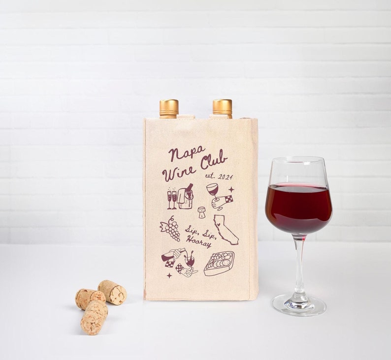 May include: A beige canvas wine tote bag with two wine bottles inside. The bag has a red and white design with the text "Napa Wine Club est. 2021" and illustrations of wine glasses, grapes, and a California map.