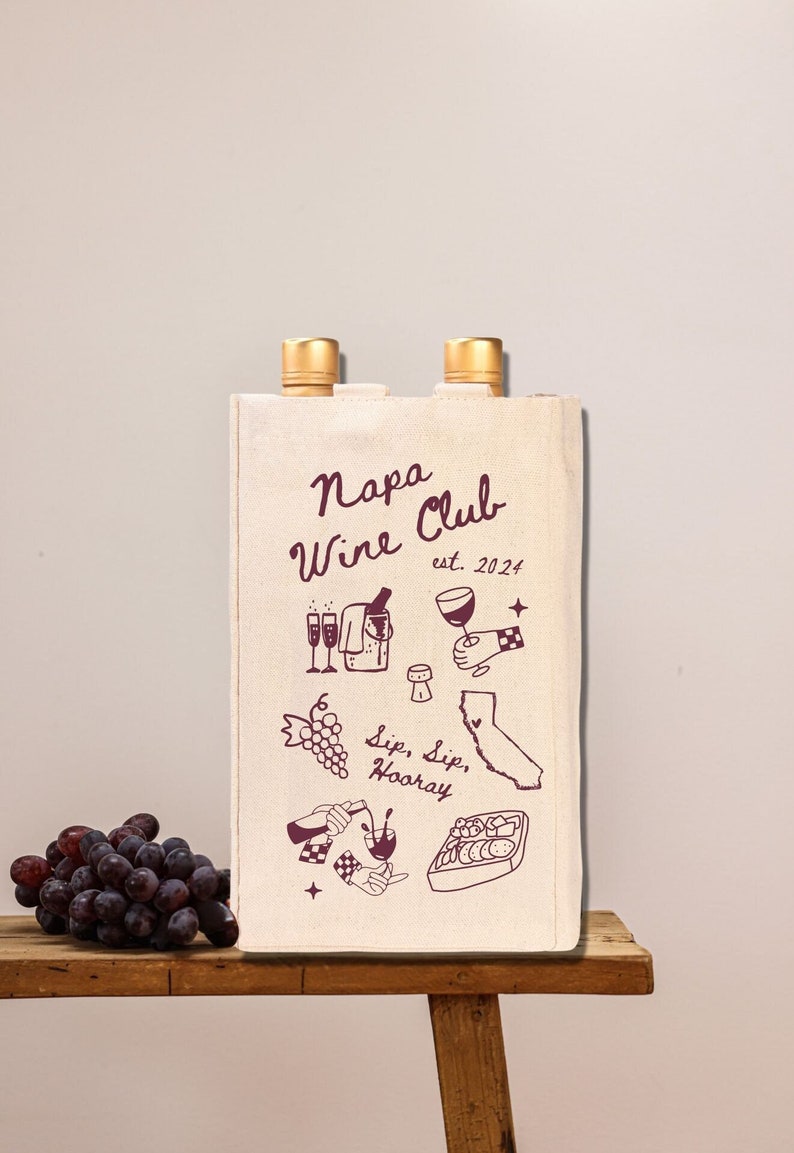May include: A white canvas wine tote bag with the text "Napa Wine Club est. 2024" and illustrations of wine bottles, grapes, glasses, and a map of California. The bag has two pockets on the sides for wine bottles.