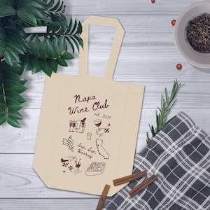 May include: A beige tote bag with a brown handle and a design that says "Napa Wine Club est. 2021" with illustrations of wine glasses, grapes, and a bottle of wine.