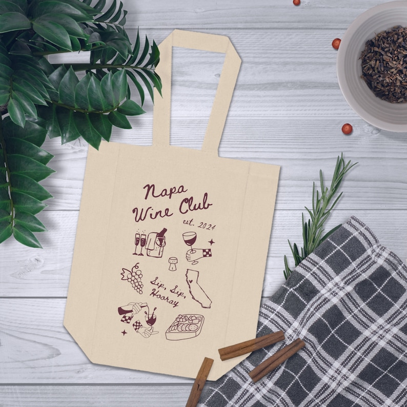 May include: A beige tote bag with a brown handle and a design that says "Napa Wine Club est. 2021" with illustrations of wine glasses, grapes, and a bottle of wine.