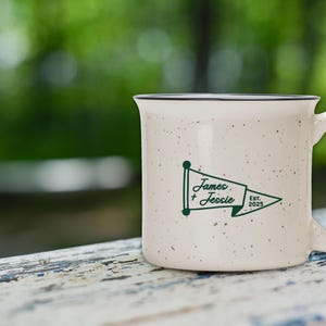 May include: Cream-colored ceramic mug with a speckled finish and a black rim. The mug features a green pennant design with the names "James + Jessie" and the year "2025". The mug is set against a blurred green background.