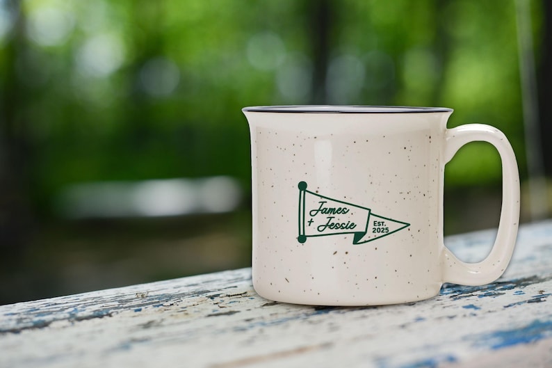 May include: Cream-colored ceramic mug with a speckled finish and a black rim. The mug features a green pennant design with the names "James + Jessie" and the year "2025". The mug is set against a blurred green background.