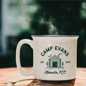 May include: A speckled white ceramic mug with the text "CAMP EVANS" and a cabin graphic in green. The mug also features the text "EST. 2025" and "Asheville, NC". Steam rises from the mug, suggesting a hot beverage.