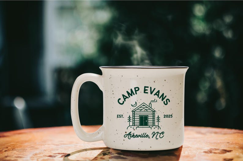 May include: A speckled white ceramic mug with the text "CAMP EVANS" and a cabin graphic in green. The mug also features the text "EST. 2025" and "Asheville, NC". Steam rises from the mug, suggesting a hot beverage.
