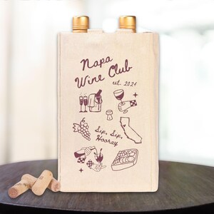 May include: A beige canvas wine tote bag with the text "Napa Wine Club est. 2024" printed in maroon. The bag features illustrations of wine bottles, glasses, grapes, and a map of California.
