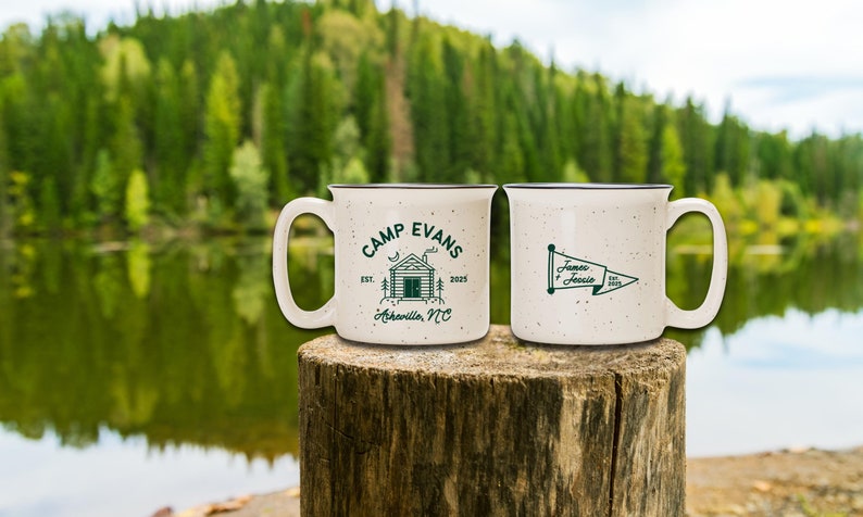 May include: Two speckled ceramic mugs with dark green designs. One mug features "Camp Evans" text with a cabin graphic and "Asheville, NC" text. The other mug has a pennant design with "James & Jessie" text. Both mugs have a dark brown rim.