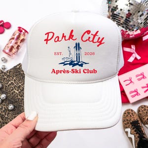 May include: White trucker hat with a mesh back and a curved brim. The front features the text "Park City" in red script, with a graphic of skis and the words "Aprés-Ski Club". The hat is part of a ski-themed collection.