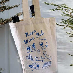 May include: A beige canvas tote bag with blue text and illustrations. The text reads "Napa Wine Club" and "Loosanky". The illustrations include wine glasses, grapes, a bottle of wine, a cork, and a map of California.