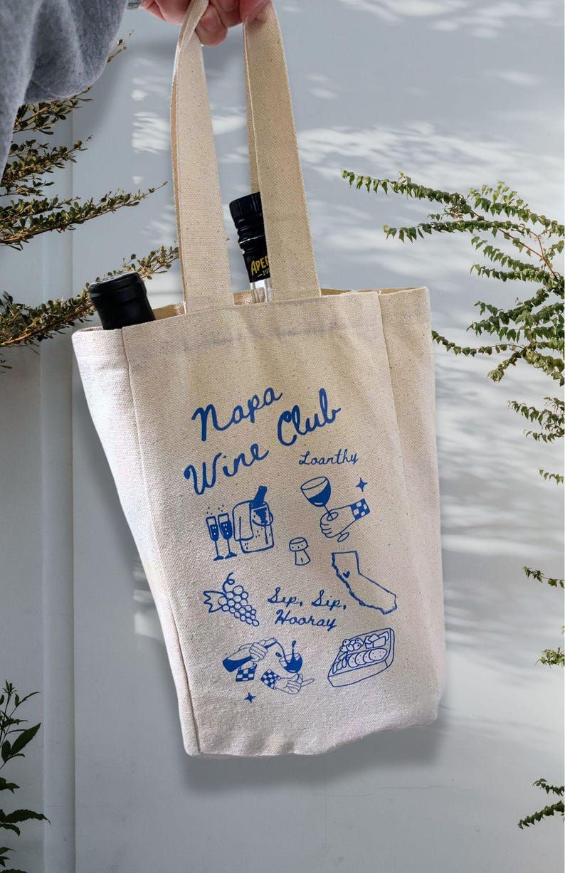 May include: A beige canvas tote bag with blue text and illustrations. The text reads "Napa Wine Club" and "Loosanky". The illustrations include wine glasses, grapes, a bottle of wine, a cork, and a map of California.