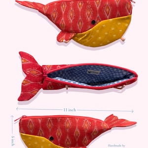 以下が含まれることがあります： 長さ28cm、高さ13cmのクジラの形をしたペンケース。外側はオレンジ色のひし形模様が入った赤い生地で、お腹部分は黄色です。内側は青と白のチェック柄の生地で裏打ちされています。