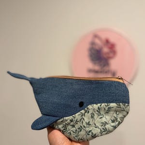 以下が含まれることがあります： 青いデニム生地で作られたクジラの形をしたポーチで、ベージュのファスナーが付いています。下部はダークグリーンの葉模様が入ったライトグリーンの生地です。小さな黒い丸いボタンがクジラの目として付いています。