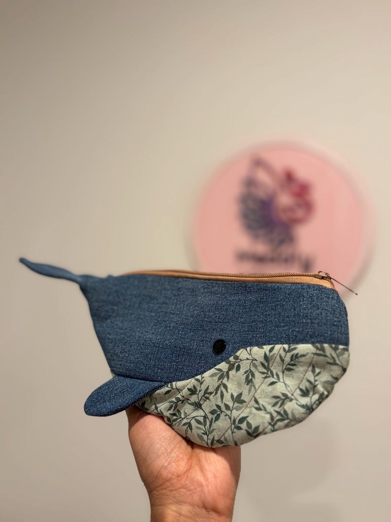 以下が含まれることがあります： 青いデニム生地で作られたクジラの形をしたポーチで、ベージュのファスナーが付いています。下部はダークグリーンの葉模様が入ったライトグリーンの生地です。小さな黒い丸いボタンがクジラの目として付いています。