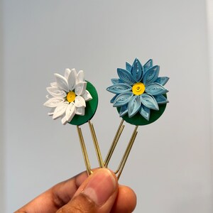 以下が含まれることがあります： 緑色の芯と金色のクリップが付いた、青い紙の花と白い紙の花のブックマーク2つ