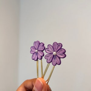 以下が含まれることがあります： 紫色の紙の花がついた金色のクリップが2つ。それぞれの花の中央には小さな真珠が埋め込まれています。