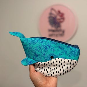 以下が含まれることがあります： クジラの形をした布製のポーチ。上部はターコイズブルー地にダークブルーの花柄、下部は白地に黒いダッシュ模様が施されています。上部にはダークブルーのファスナーがあり、小さな黒いボタンの目が付いています。
