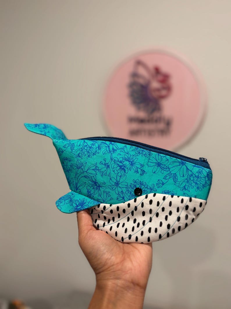 以下が含まれることがあります： クジラの形をした布製のポーチ。上部はターコイズブルー地にダークブルーの花柄、下部は白地に黒いダッシュ模様が施されています。上部にはダークブルーのファスナーがあり、小さな黒いボタンの目が付いています。
