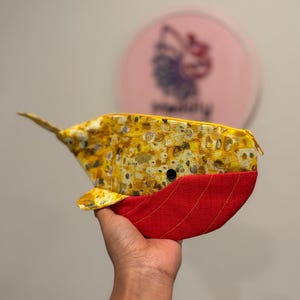 以下が含まれることがあります： 黄色と茶色のまだら模様の上半分と、赤色の質感のある生地の下半分で構成された、クジラの形をした布製ポーチ。ポーチには小さな黒い円形の目と、上部に黄色のファスナーが付いています。