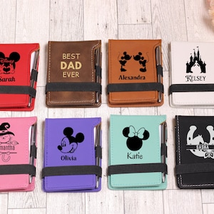 May include: Eight personalized leatherette notebooks with a pen attached. Each notebook features a different Disney character or theme, including Mickey Mouse, Minnie Mouse, a castle, a horseshoe, a witch, and a heart. The notebooks are in various colors, including red, brown, white, purple, blue, black, and pink.