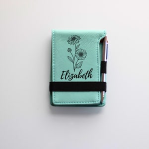 May include: A light blue leather notebook with a black elastic band and a silver pen. The notebook has a floral design and the name "Elizabeth" is printed on the front.