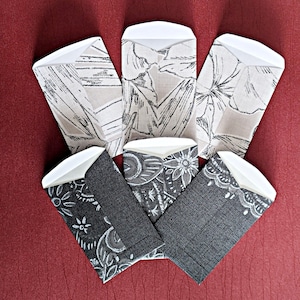 Könnte beinhalten: Sechs kleine Umschläge mit verschiedenen Mustern. Die Umschläge sind weiß mit schwarzen und grauen Designs. Einige der Umschläge haben Blumenmuster, während andere geometrische Muster haben.