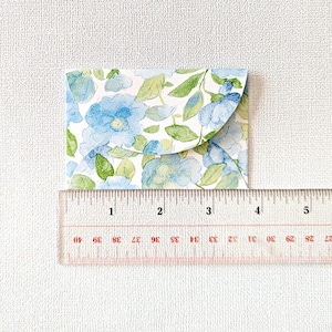 Puede incluir: Un pequeño sobre con estampado floral con un diseño de acuarela azul y verde. El sobre mide aproximadamente 5 cm de ancho, con una regla en primer plano para la escala. El diseño presenta flores azules y hojas verdes sobre un fondo blanco.