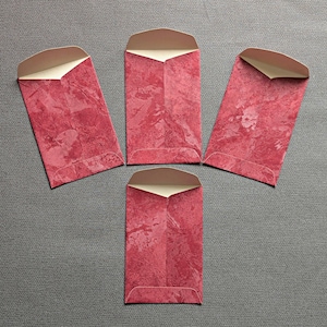 May include: Four red envelopes with a marbled texture and a cream-colored interior. The envelopes are rectangular with a curved top flap. The envelopes are arranged on a gray surface.