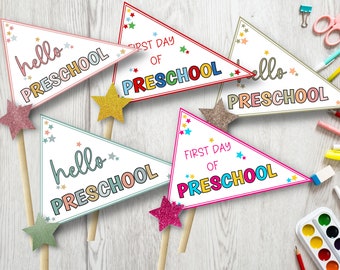 Op de afbeelding: Vier kleurrijke driehoekige vlaggetjes met de tekst "hello preschool" en "first day of preschool" op een witte achtergrond met glittersterren. De vlaggetjes zijn bevestigd aan houten stokjes.
