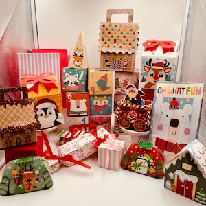 May include: A collection of festive Christmas-themed gift boxes and containers. The assortment includes boxes shaped like houses, sweaters, and cubes, decorated with reindeer, penguins, and other holiday characters. Some boxes have the text "OH WHAT FUN".