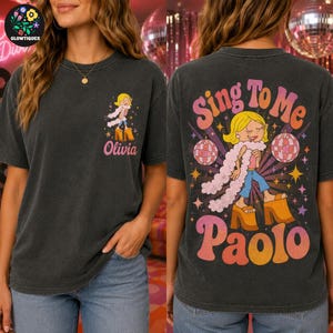 May include: A charcoal gray t-shirt featuring a graphic of a blonde character with a pink boa and platform shoes. The front displays the name Olivia, while the back shows the same character with disco balls and the text Sing To Me Paolo.
