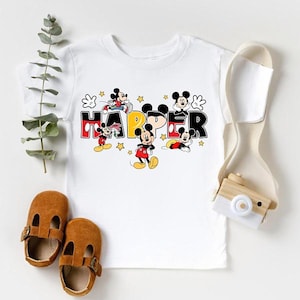 A white t-shirt with a Mickey Mouse design, a pair of brown baby shoes, and a camera are arranged on a white background.