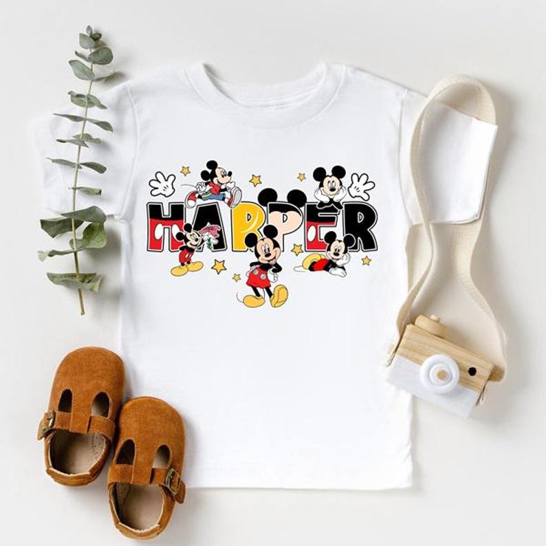 A white t-shirt with a Mickey Mouse design, a pair of brown baby shoes, and a camera are arranged on a white background.