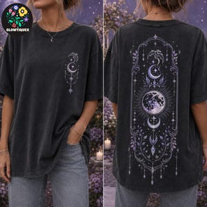 May include: A charcoal gray short-sleeve t-shirt featuring a small celestial graphic on the front left chest and a large, intricate vertical design on the back. The back print displays a moon, stars, and floral elements in shades of purple and white.