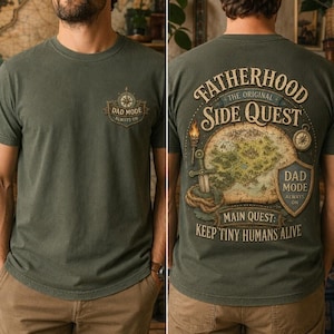 May include: An olive green t-shirt featuring a small compass graphic on the front chest with the text DAD MODE ALWAYS ON. The back displays a large fantasy map illustration with a sword, shield, and the text FATHERHOOD SIDE QUEST MAIN QUEST: KEEP TINY HUMANS ALIVE.